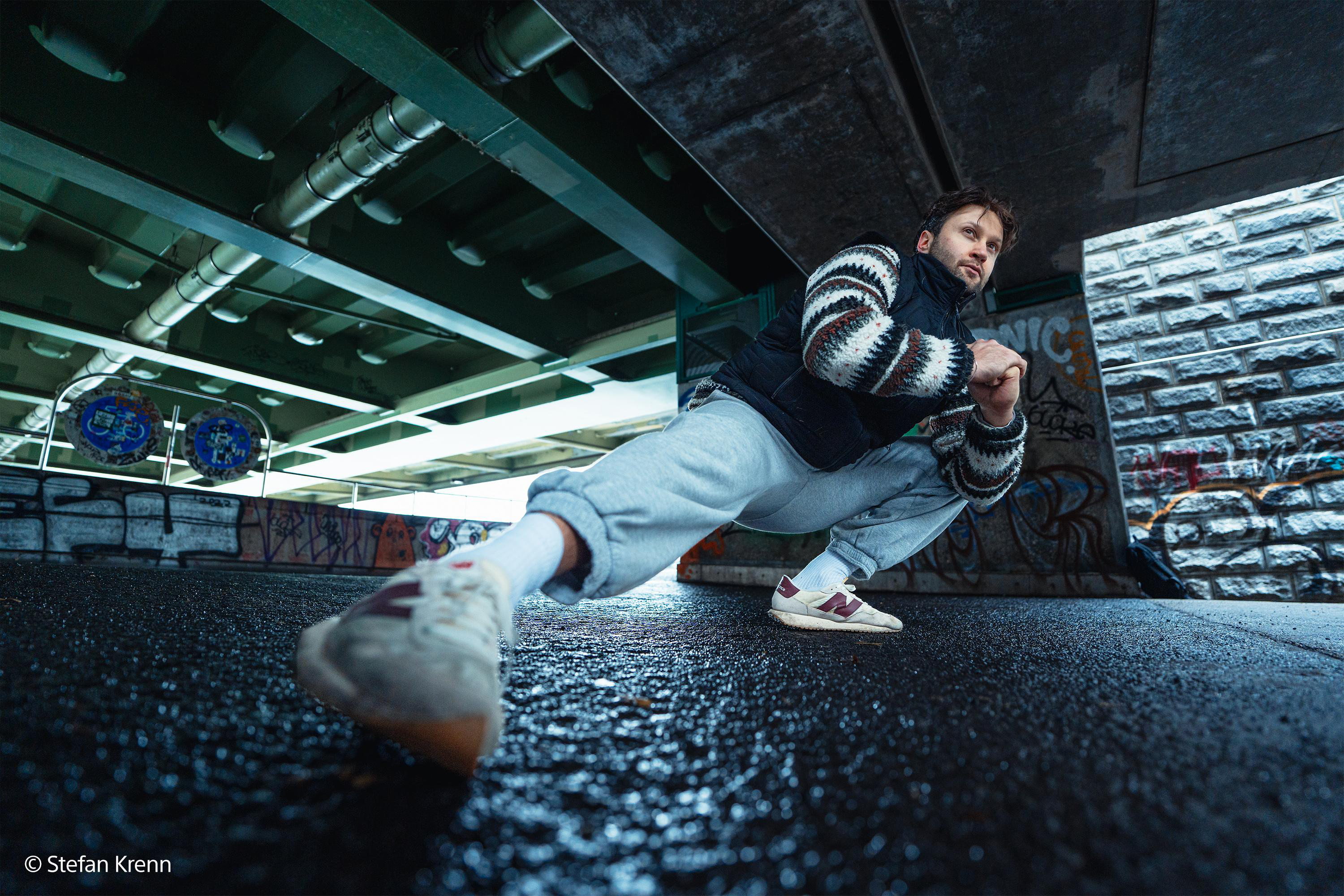 Mann macht eine tiefe Hocke unter einer Brücke mit Rohren und Graffiti.