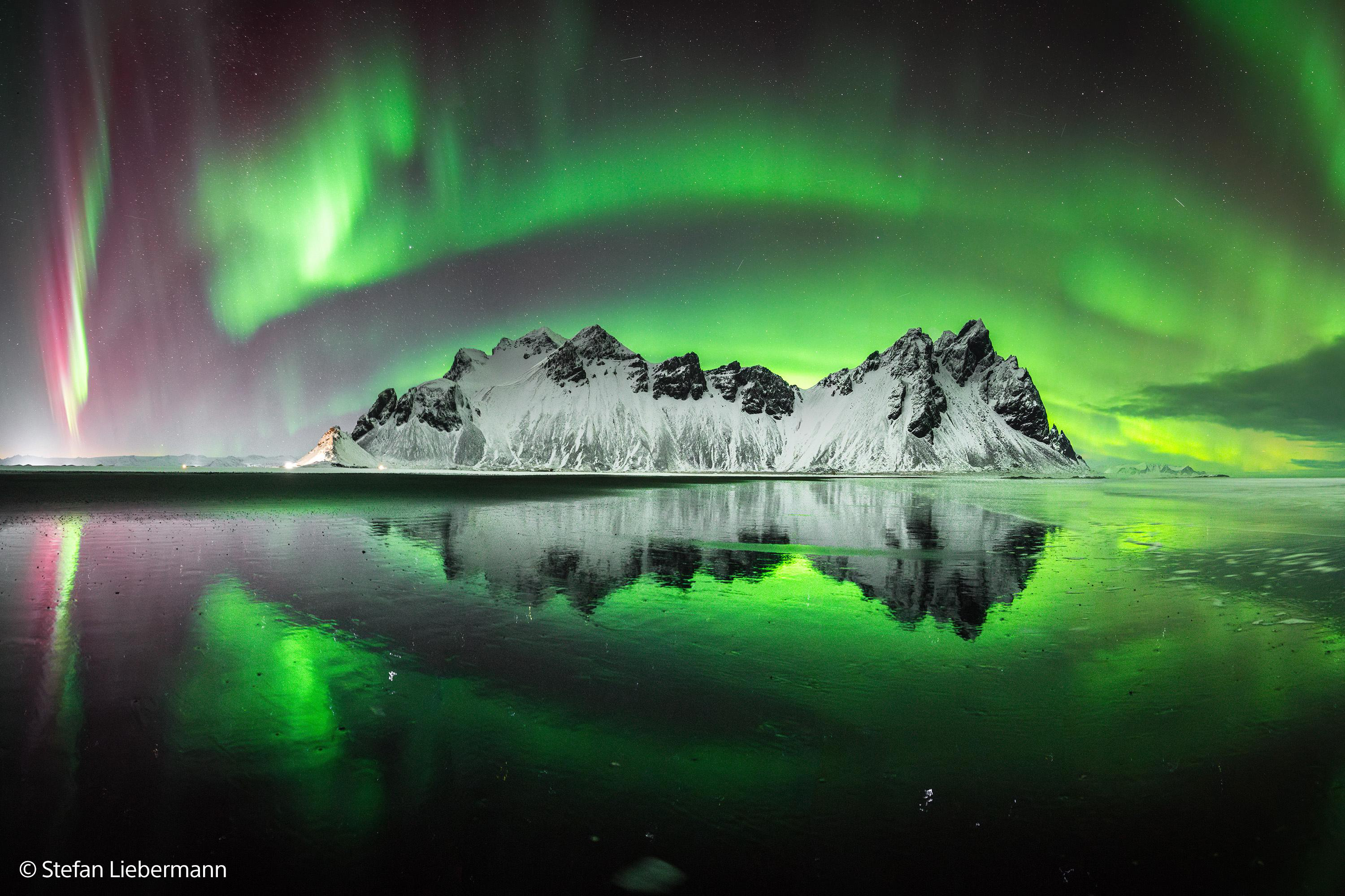 Grüne und rote Aurora über schneebedeckten Bergen, reflektiert auf einer Eisfläche.