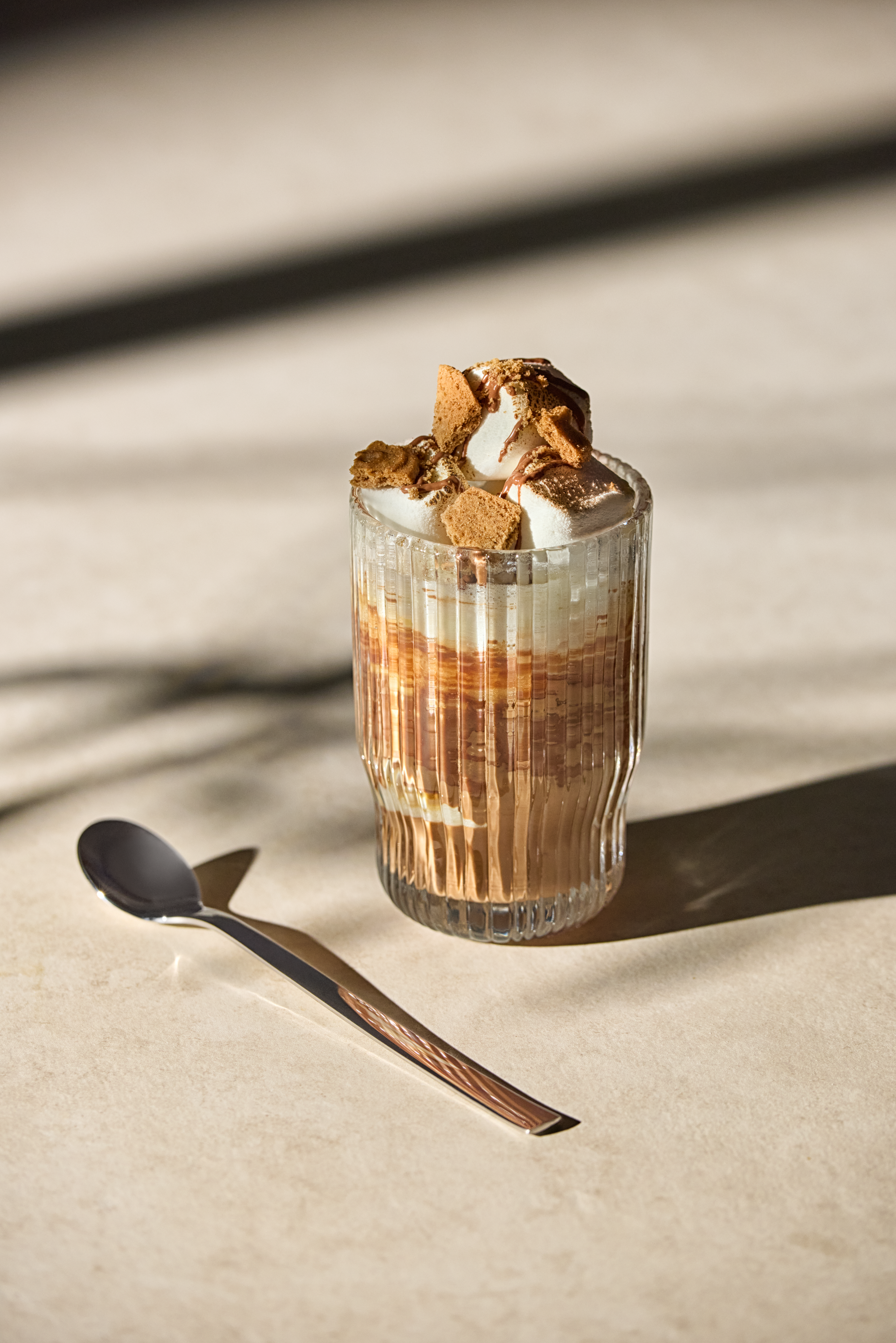 Ein Glas mit Kaffee, Sahne, Marshmallows und Keksen daneben ein Löffel.