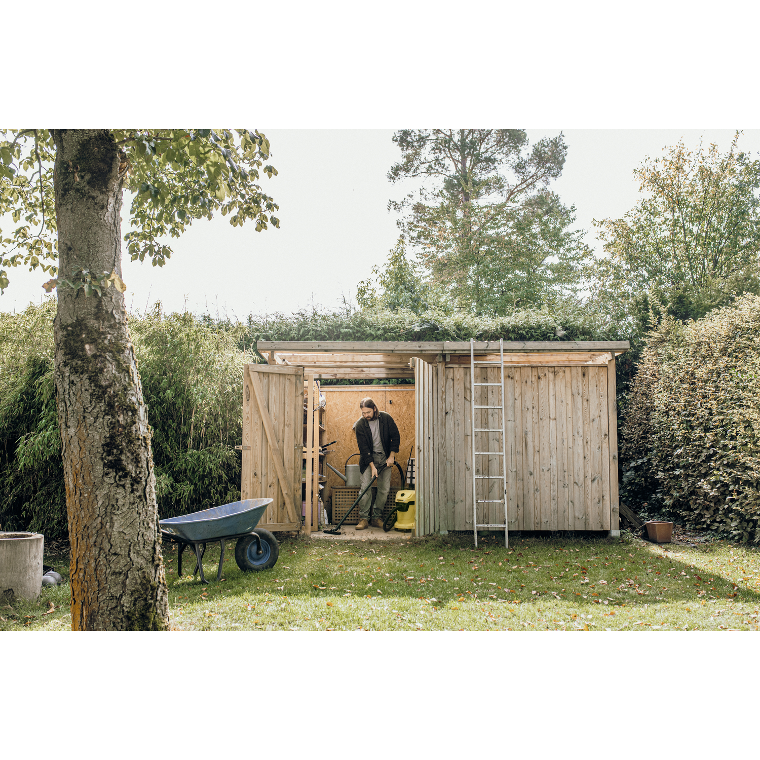 A man vacuums with a workshop vacuum cleaner outside a wooden shed.
