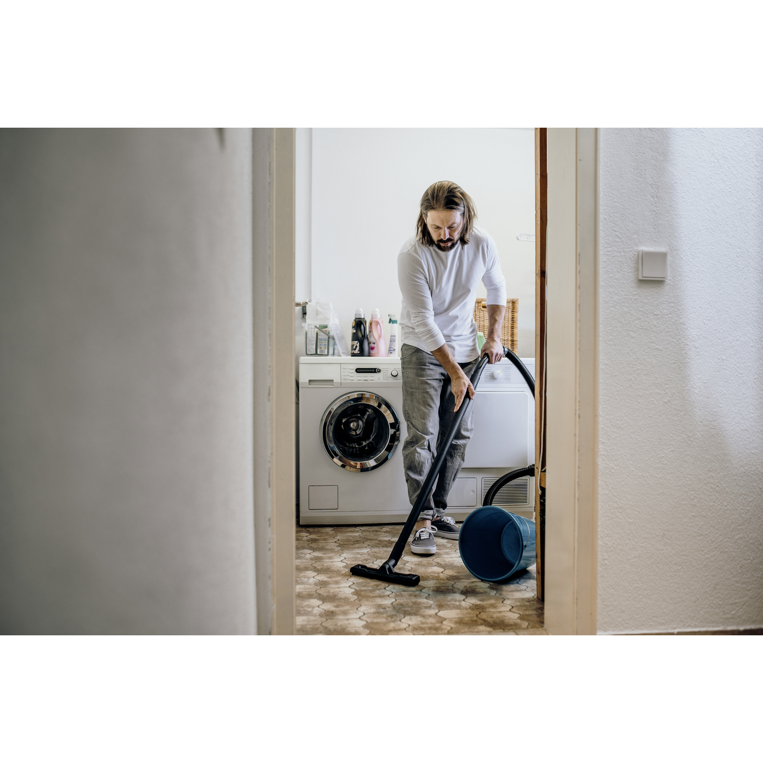 Man vacuums floor in laundry room next to washing machine and blue bin.