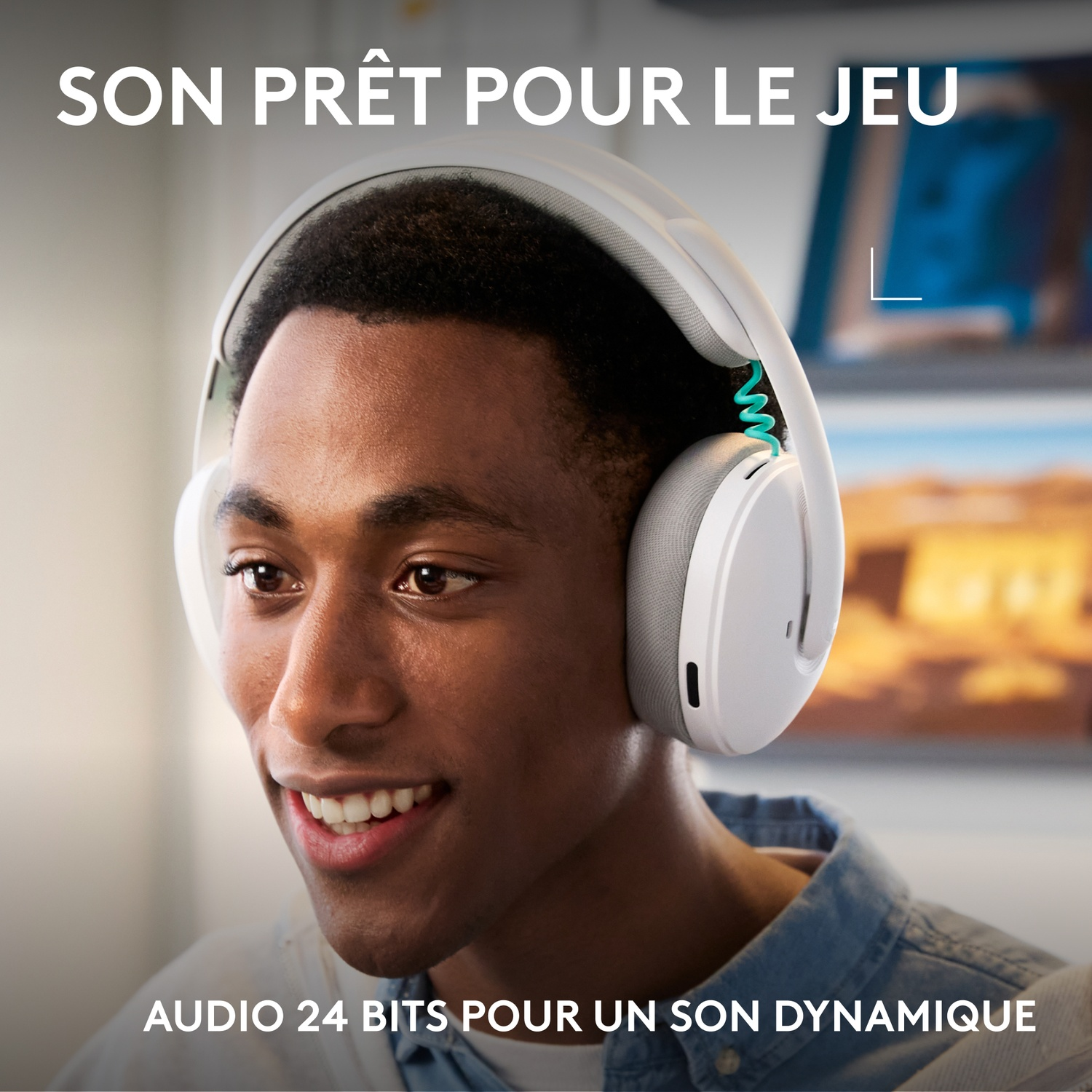 A young man wears white headphones and smiles. Screens are visible in the background.