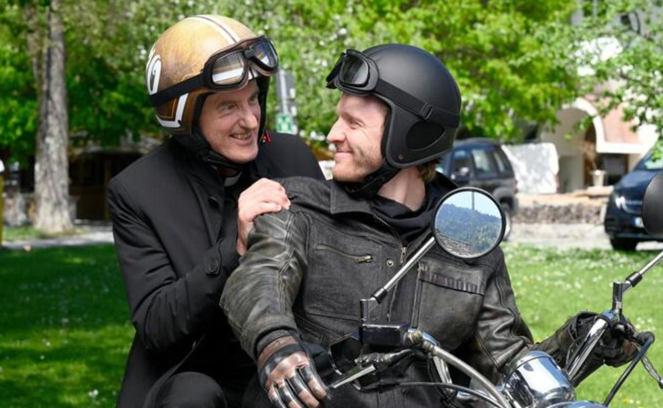 Zwei Männer auf einem Motorrad. Einer trägt einen schwarzen Anzug, der andere eine Lederjacke.