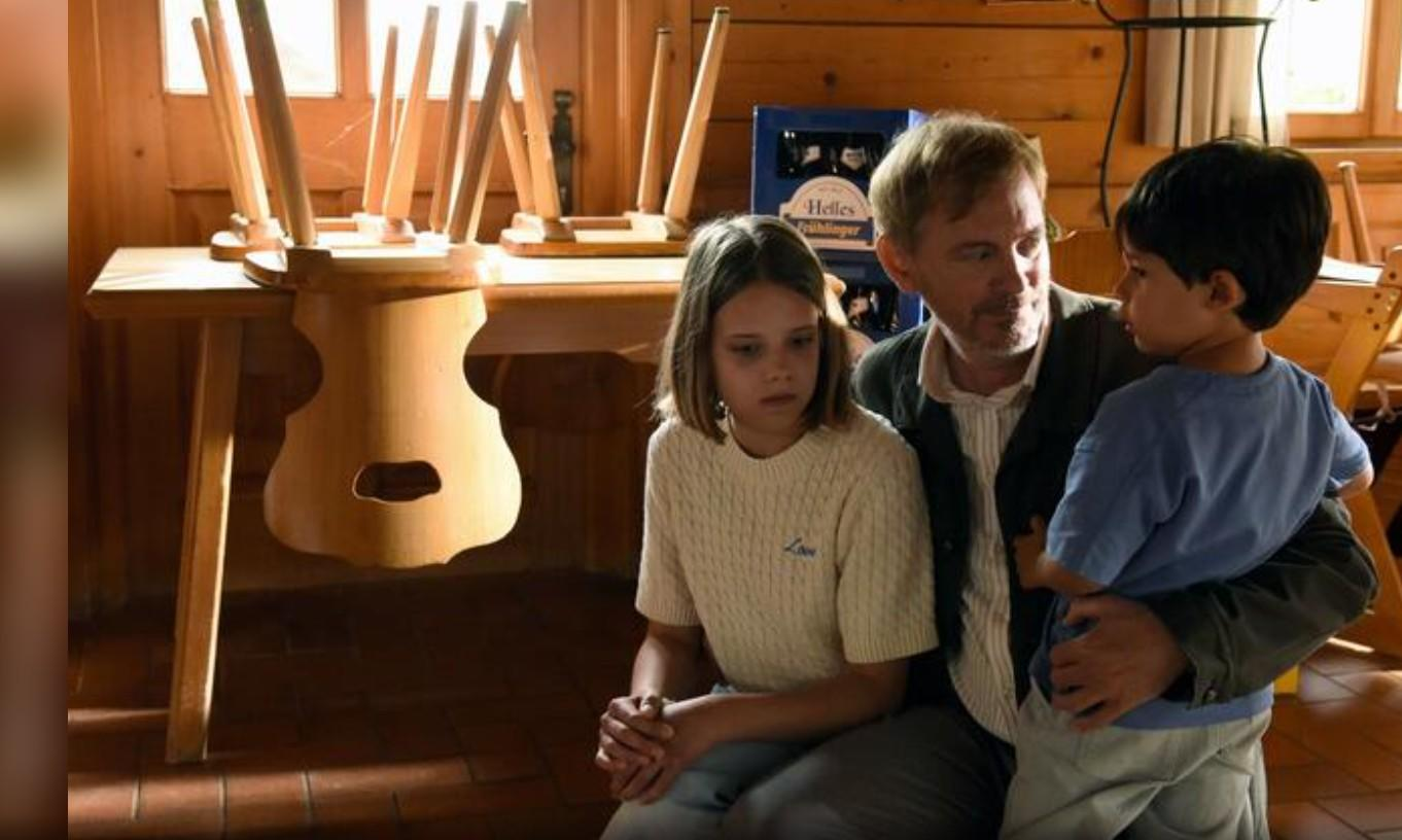 Ein Mann hält einen Jungen, ein Mädchen sitzt daneben. Tische und Stühle sind umgedreht.