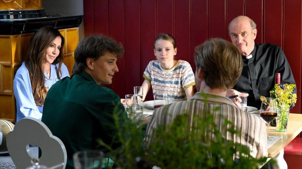 Fünf Personen sitzen an einem Tisch. Ein Priester, eine Frau, zwei junge Männer und ein Mädchen.