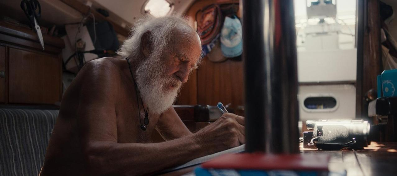 Älterer Mann mit weißem Bart schreibt auf einem Boot in ein Notizbuch.
