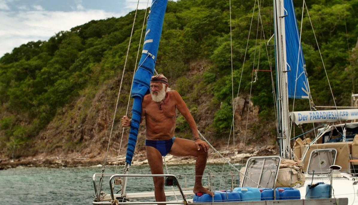 Ein älterer Mann mit Bart steht auf einem Segelboot vor einer bewaldeten Küste.