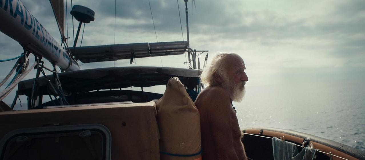 Ein älterer Mann mit weißem Haar und Bart blickt aufs Meer, auf einem Segelboot.