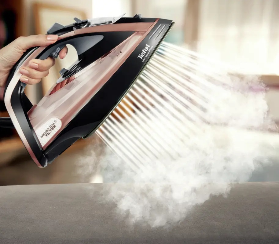 Ręka trzyma żelazko, które rozpyla parę na szarą sofę.