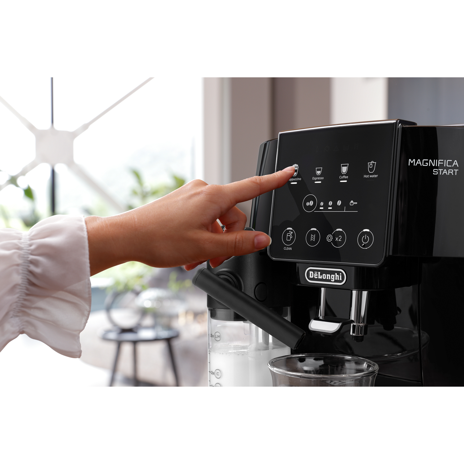 Een vinger drukt op de cappuccino-knop van een zwarte DeLonghi Magnifica Start koffiezetapparaat.