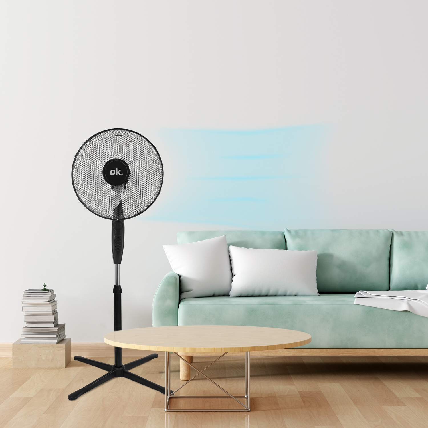 Un ventilatore a piantana nero è accanto a una pila di libri e un divano.