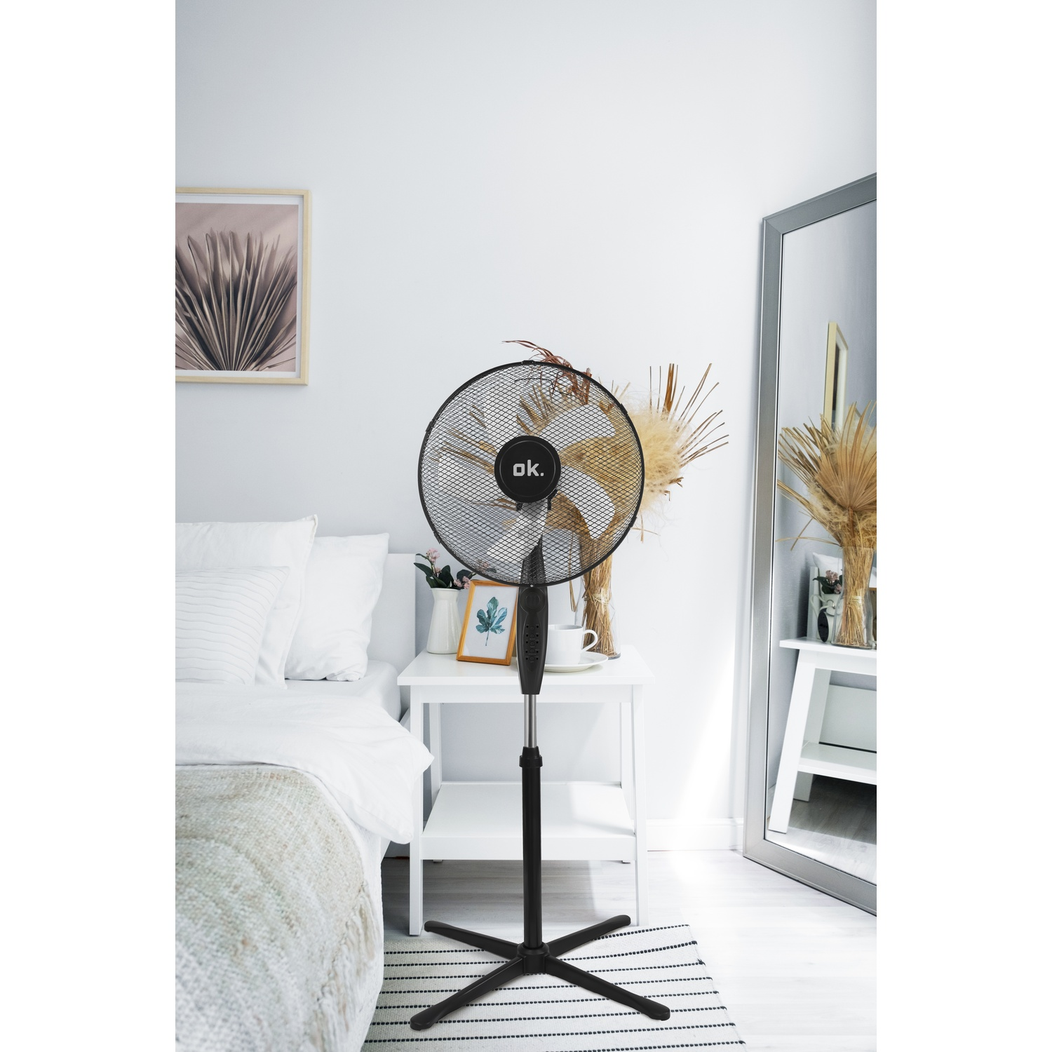 Un ventilatore nero accanto a un letto con cuscini bianchi. Decorazioni su un tavolino.