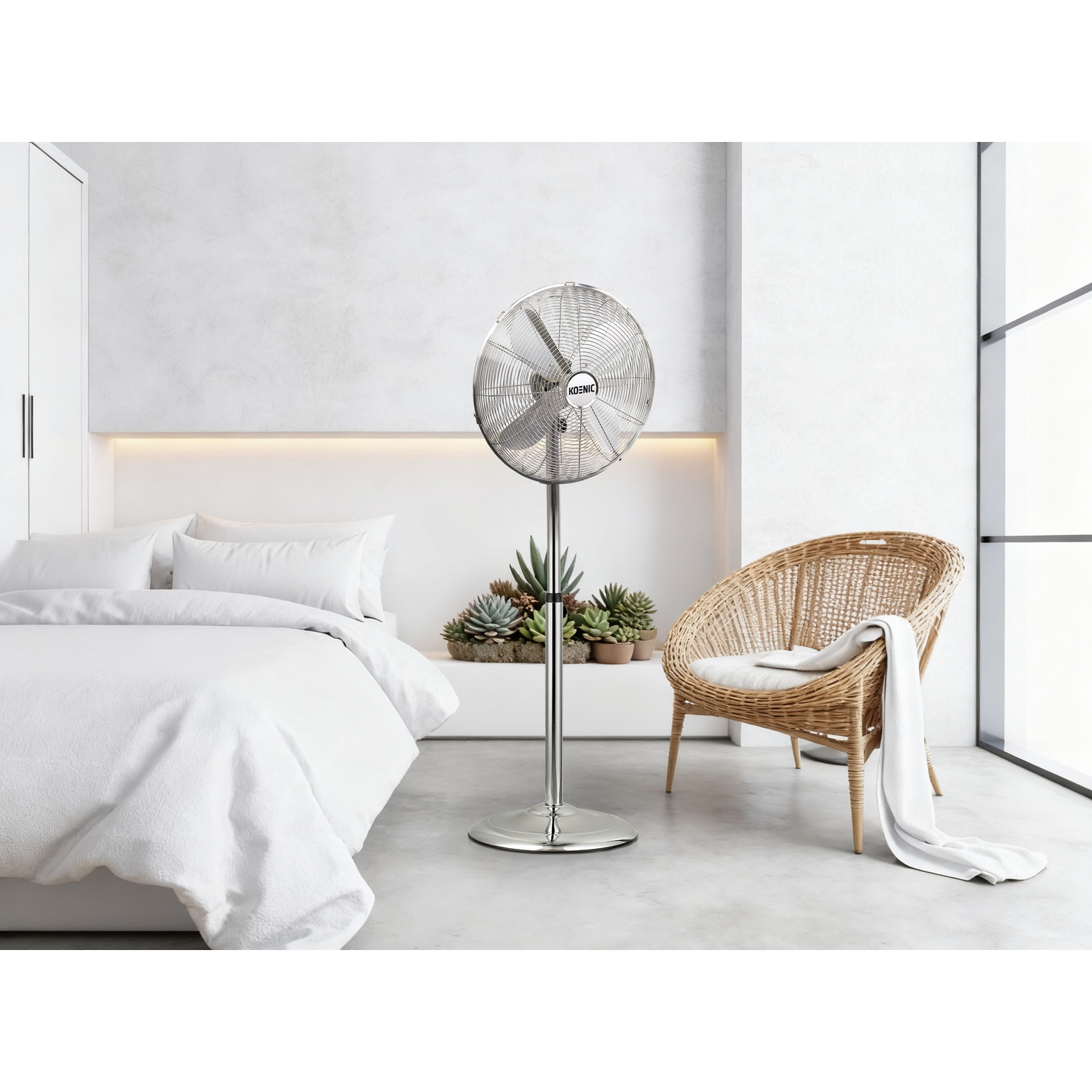 Un ventilatore da terra argentato accanto a un letto bianco e una sedia in vimini.