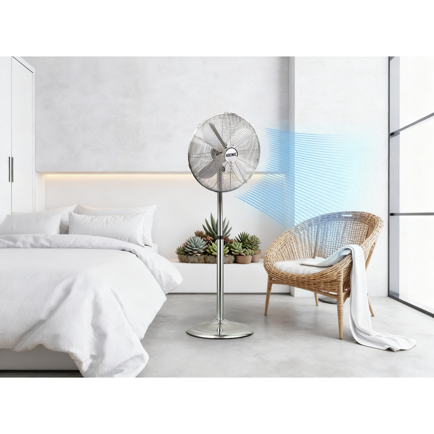 Un ventilatore da terra argentato soffia aria in una camera da letto con letto bianco e sedia in vimini.