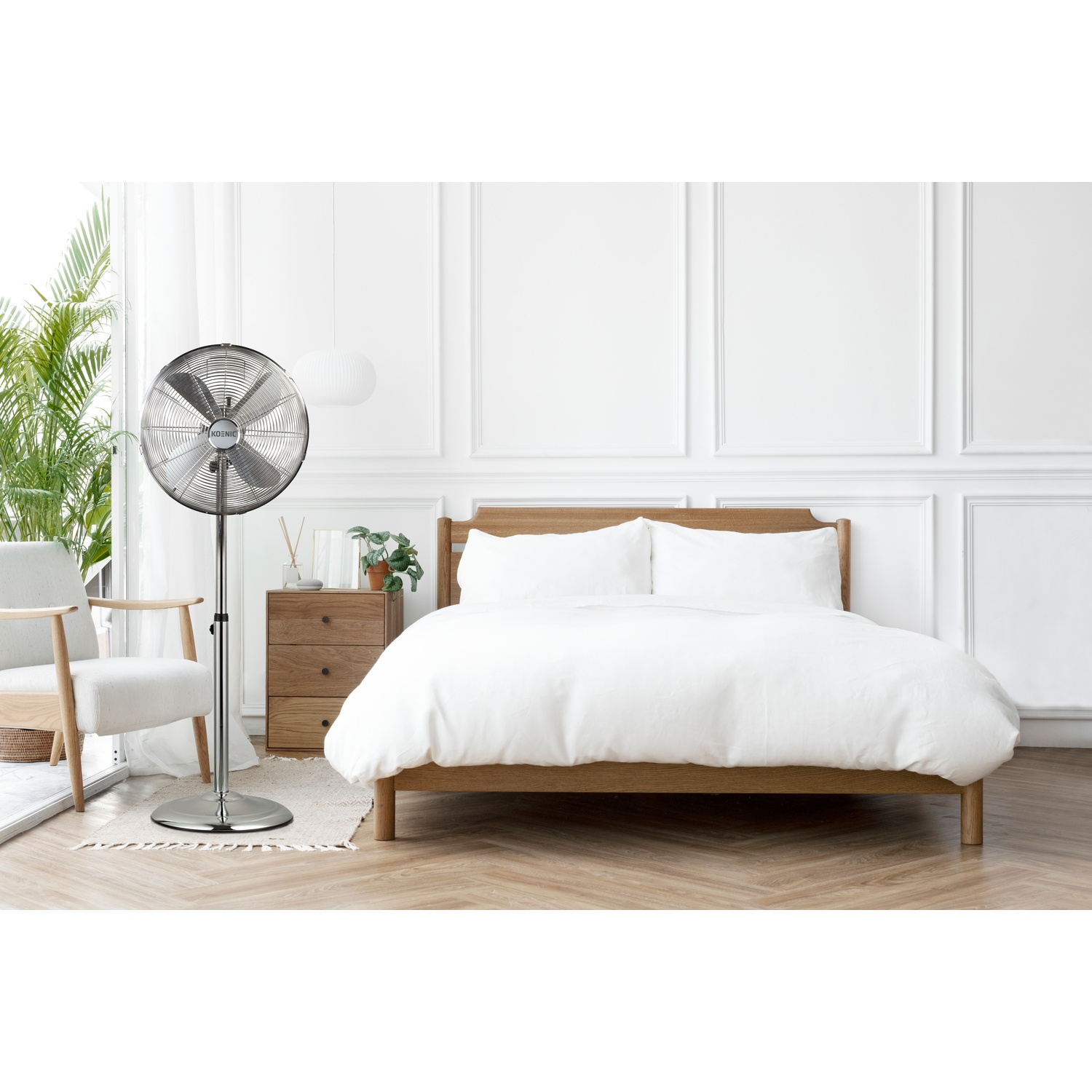 Un ventilatore da terra argentato è accanto a un letto con biancheria da letto bianca.