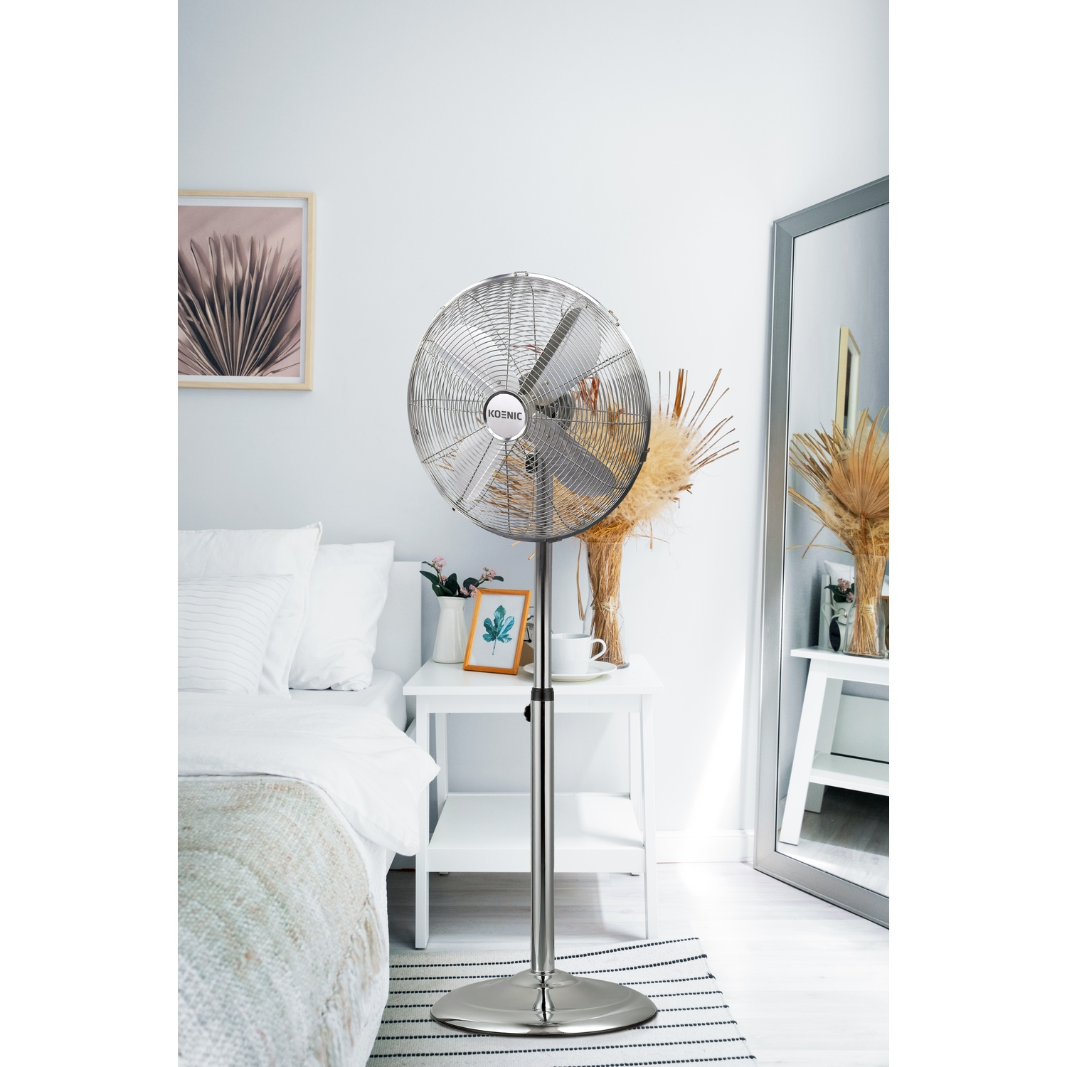 Un ventilatore da terra argentato si erge in una camera da letto accanto a un letto e un comodino.
