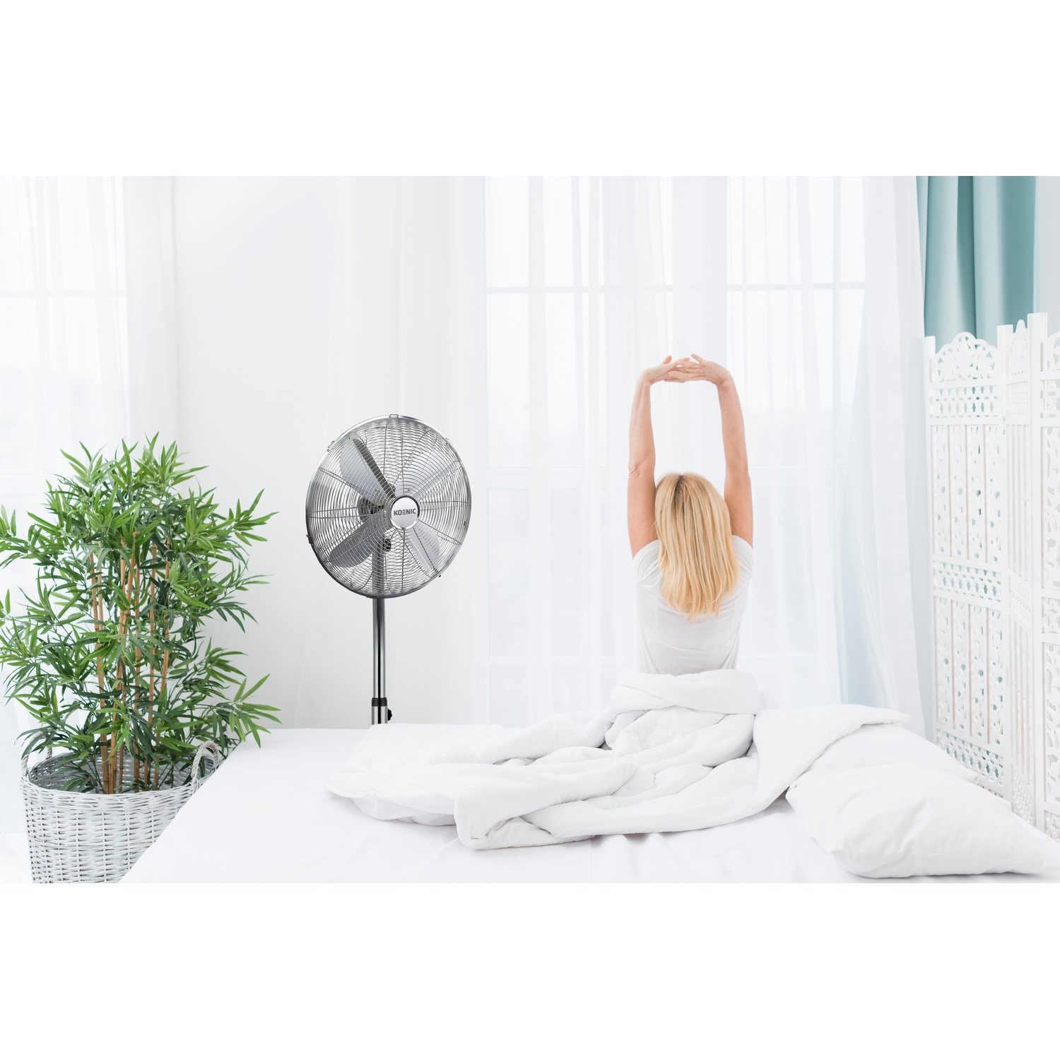 Donna si stiracchia a letto, ventilatore e pianta nella stanza.