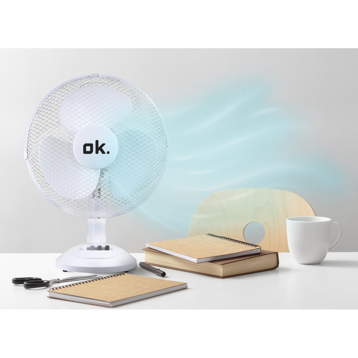 Un ventilatore bianco su una scrivania con libri, quaderni e una tazza.