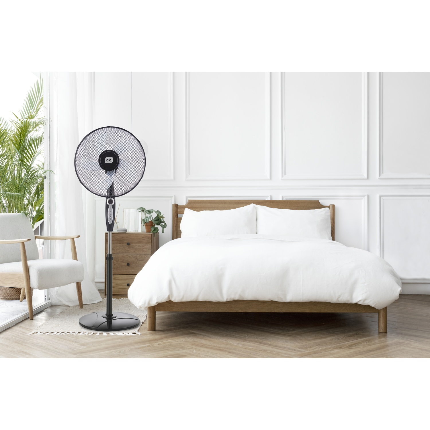 Un ventilatore oscillante nero accanto a un comodino in legno e una poltrona bianca vicino a un letto.