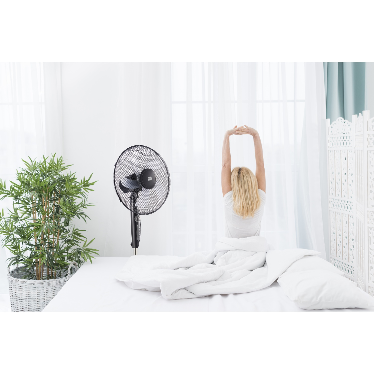 Donna che si stiracchia a letto, ventilatore e pianta vicini.