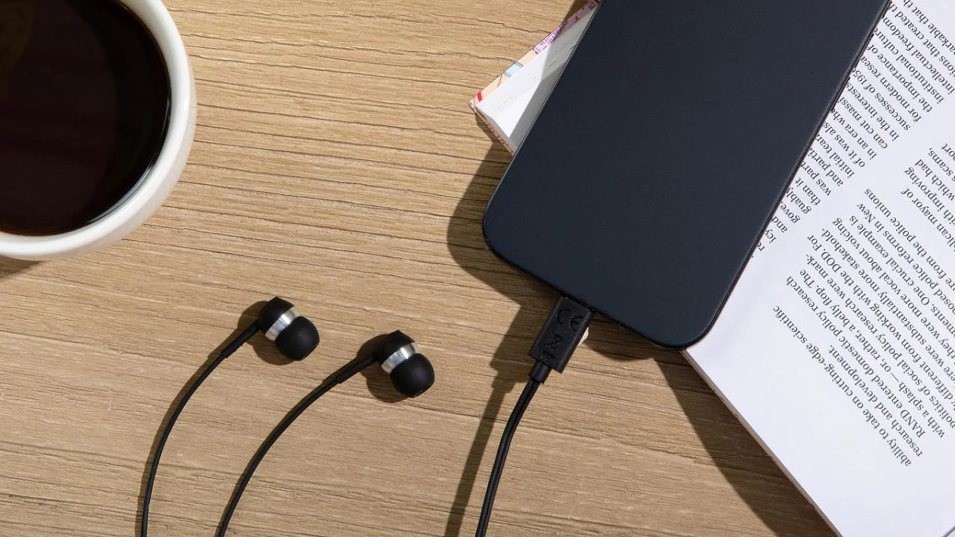 Ein schwarzes Smartphone ist mit Kopfhörern und einer Kaffeetasse auf einem Holztisch verbunden.
