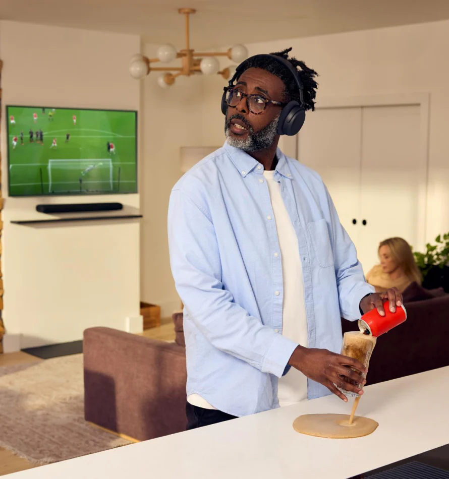 Homme avec casque verse du cola dans un verre, renversant sur le comptoir. Foot à la TV.