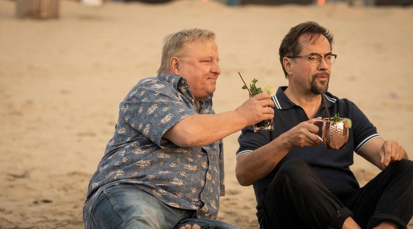 Zwei Männer sitzen am Strand und trinken Cocktails. Einer trägt ein blaues Hemd, der andere ein dunkles Poloshirt.