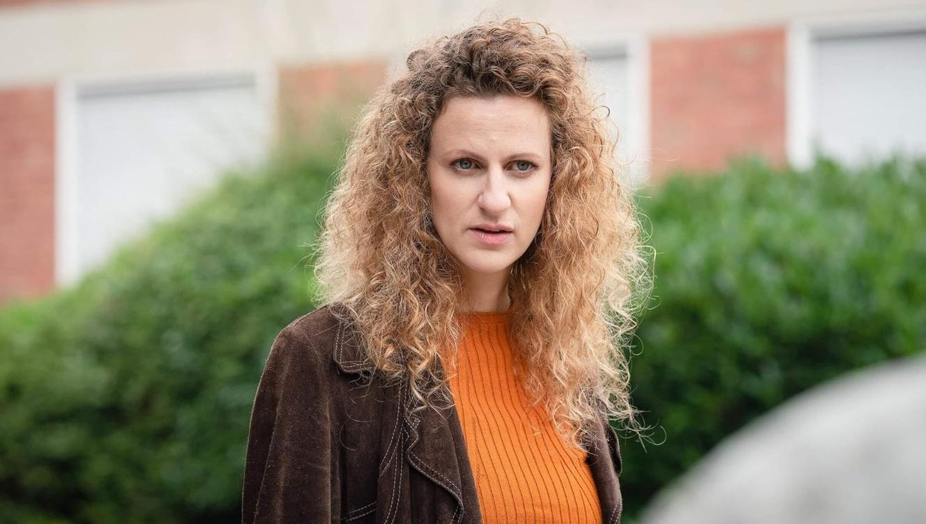 Eine Frau mit lockigen Haaren trägt eine orangefarbene Bluse und eine braune Jacke.