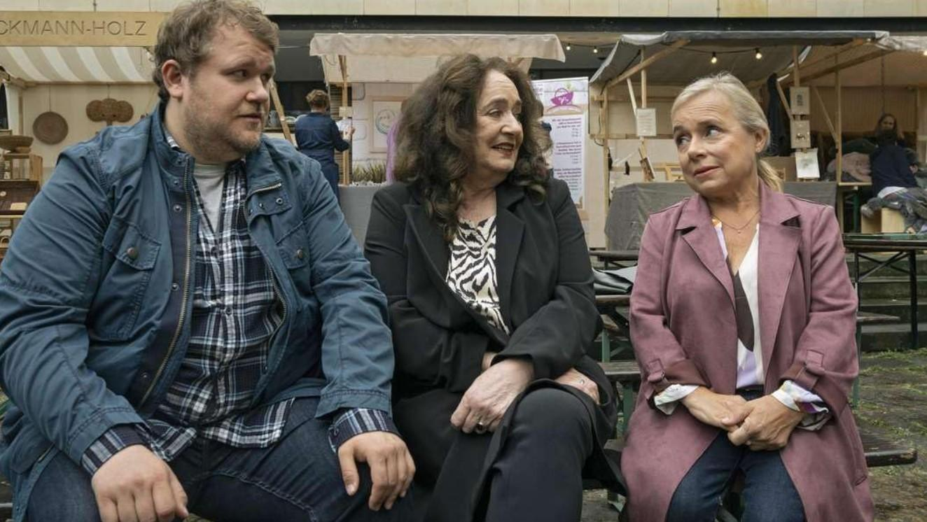 Drei Personen sitzen auf einer Bank. Ein Mann links, eine Frau Mitte, eine Frau rechts.
