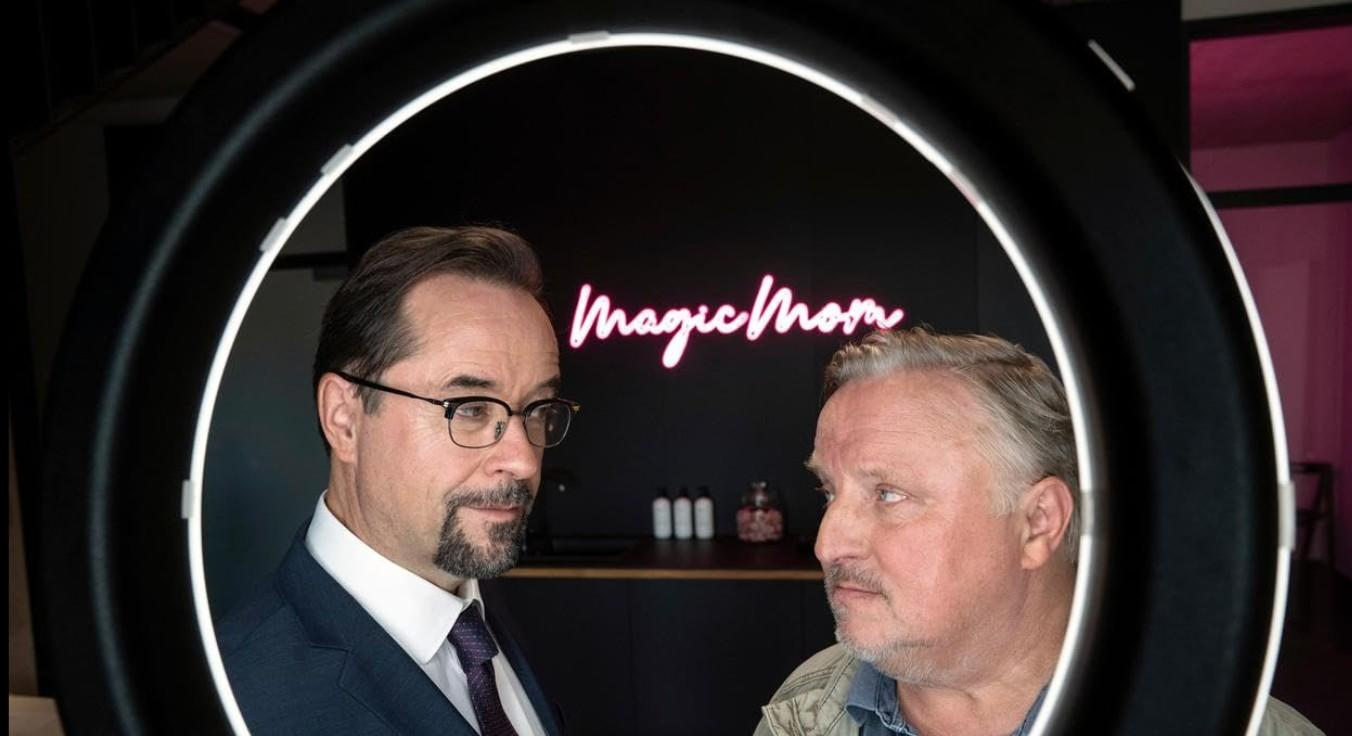 Zwei Männer blicken sich an, neonrosa Schriftzug 'Magic Mom' im Hintergrund.