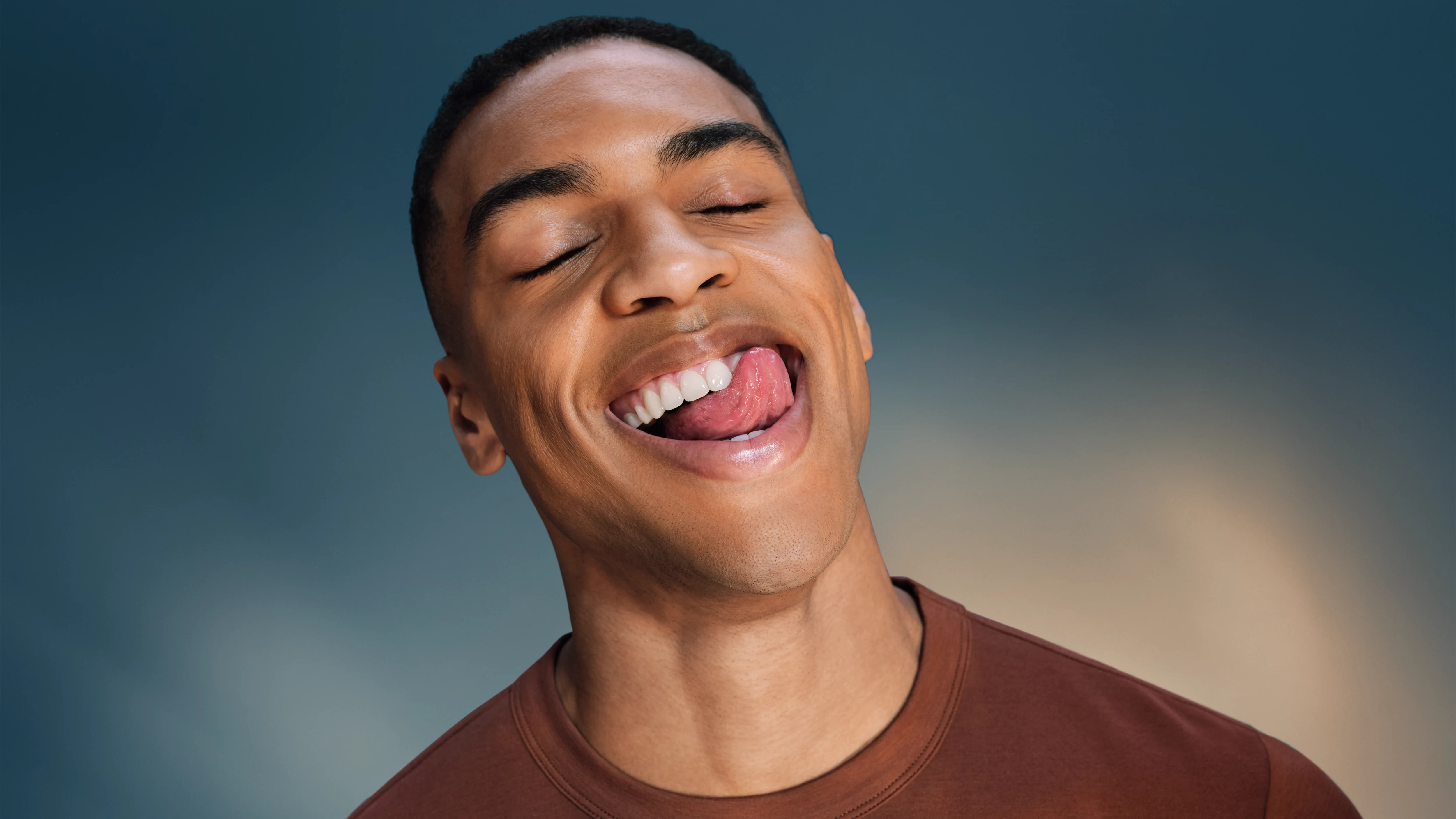 Mężczyzna z zamkniętymi oczami, oblizuje usta, nosi brązową koszulkę.