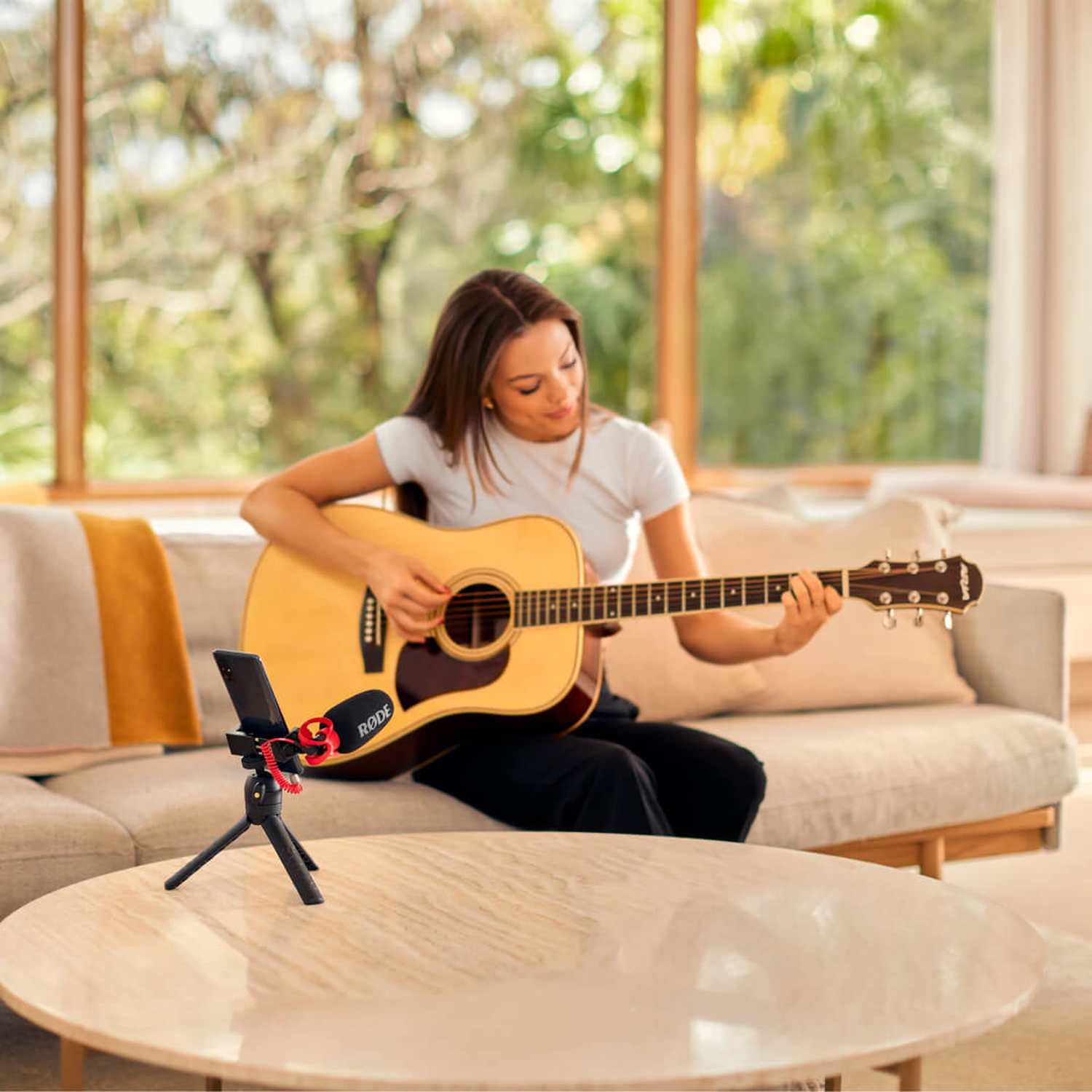 Woman plays guitar on a sofa, phone with microphone in foreground.