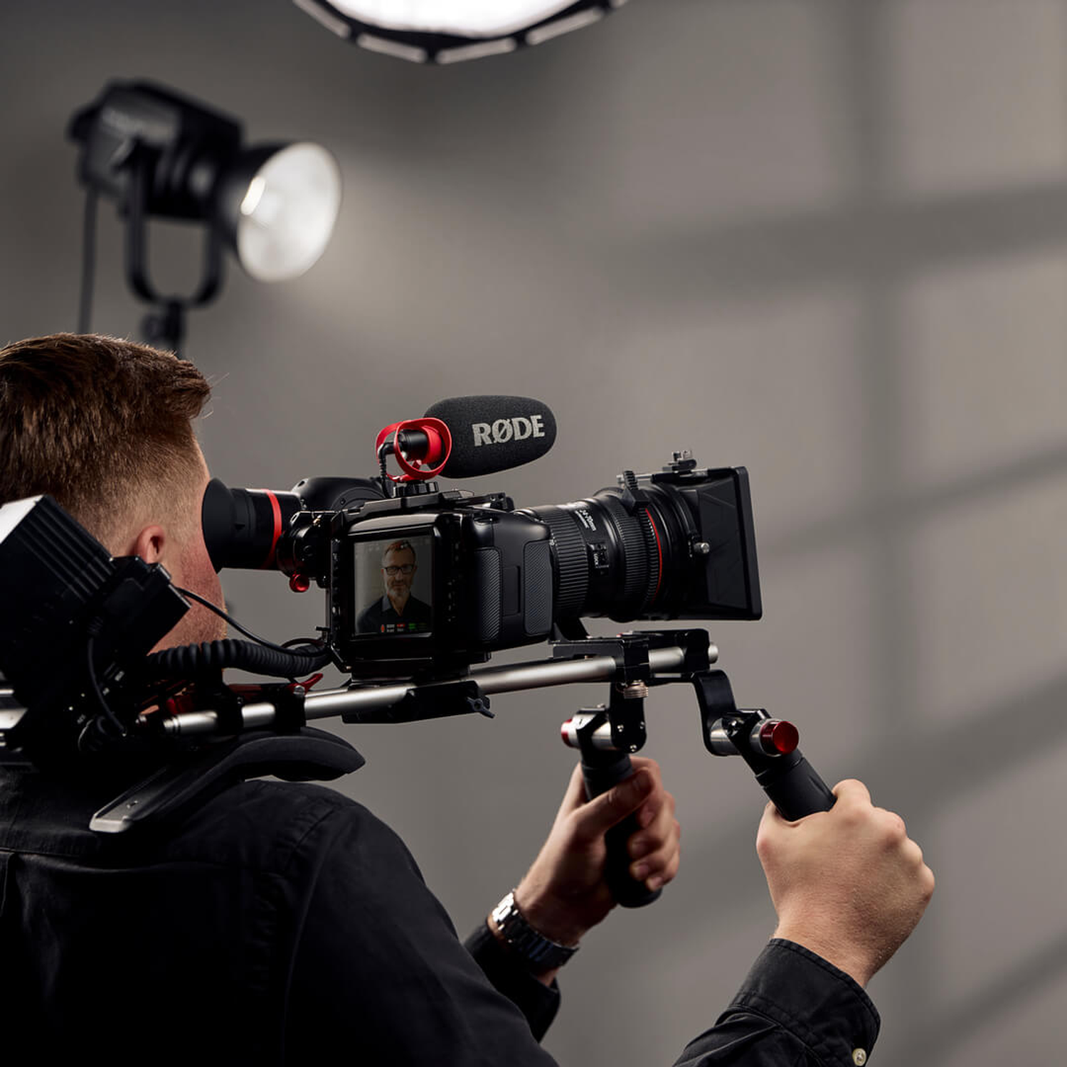 A man in black films with a shoulder-mounted camera rig, with studio lighting in background.