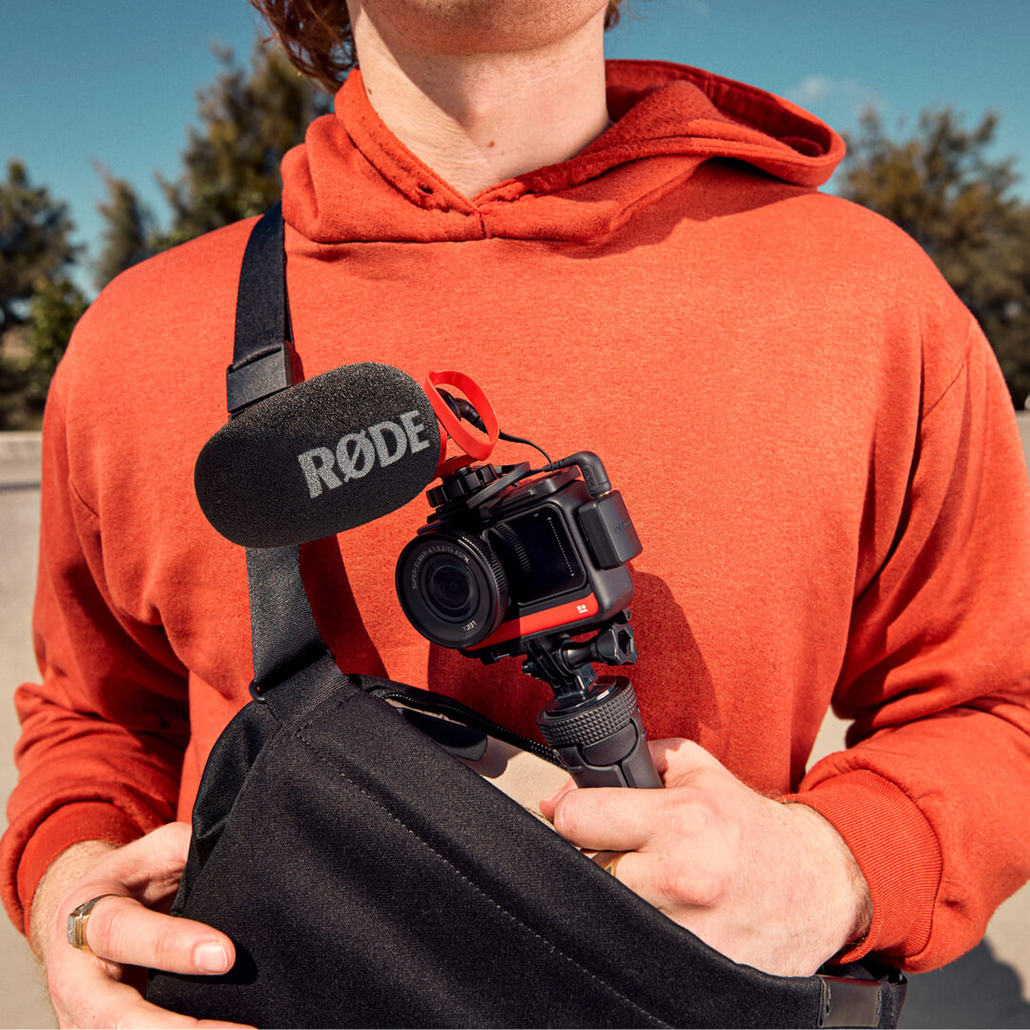 Man in orange hoodie holds camera with mic and bag. Outdoor setting.