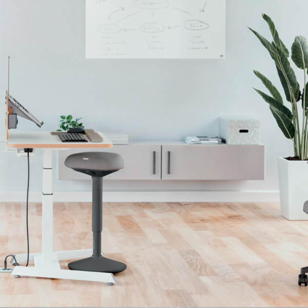 Ein Stehschreibtisch mit Laptop, Tastatur und einem Hocker in einem hellen Büro.