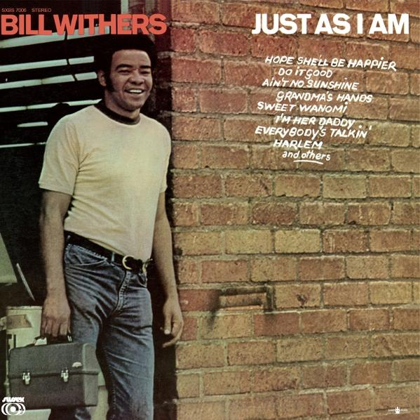 Bill Withers steht vor einer Ziegelwand, hält eine Aktentasche.