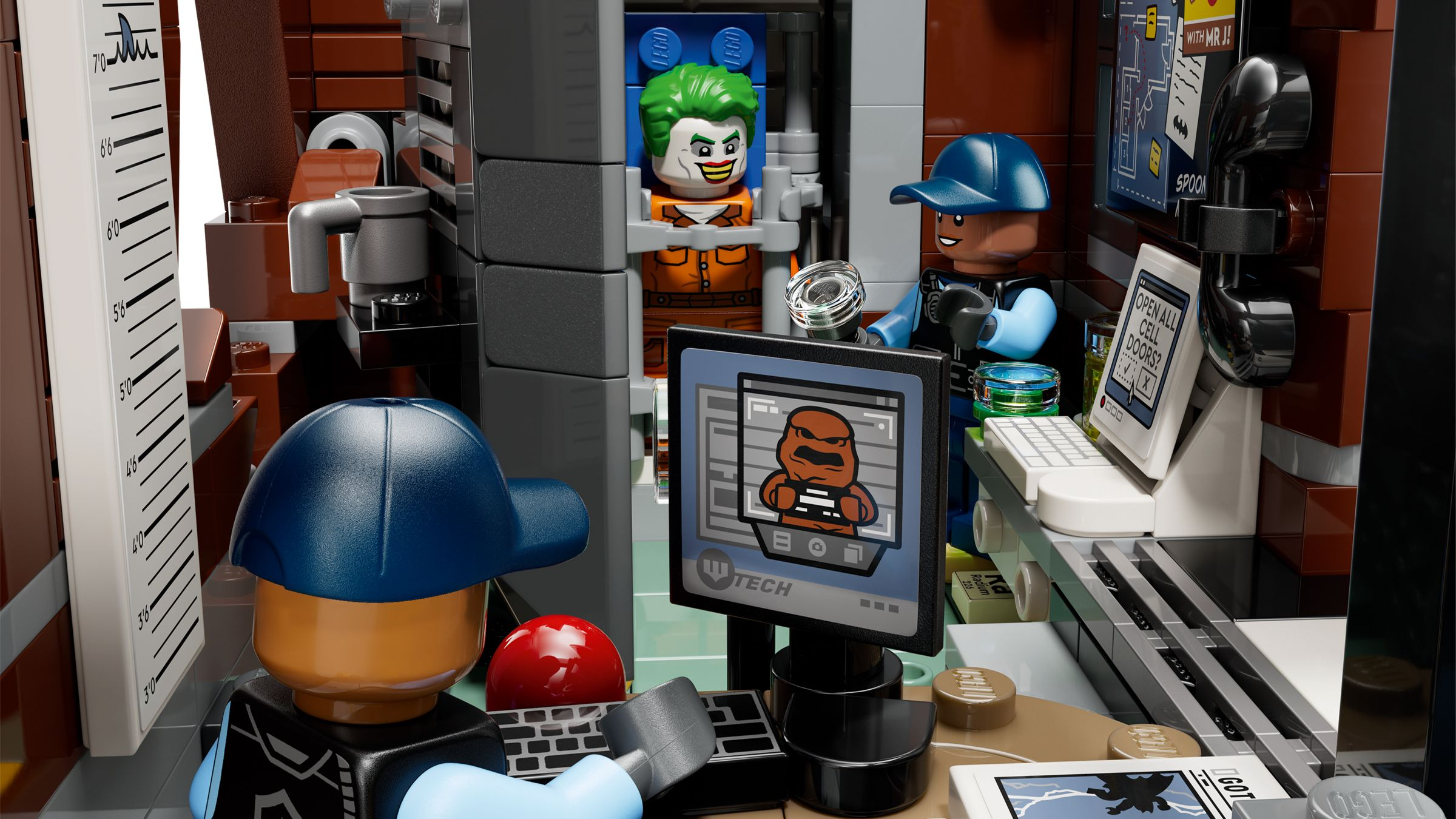 Scena więzienia Lego z Jokerem, strażnikiem przy komputerze, miarką wzrostu i telefonem.