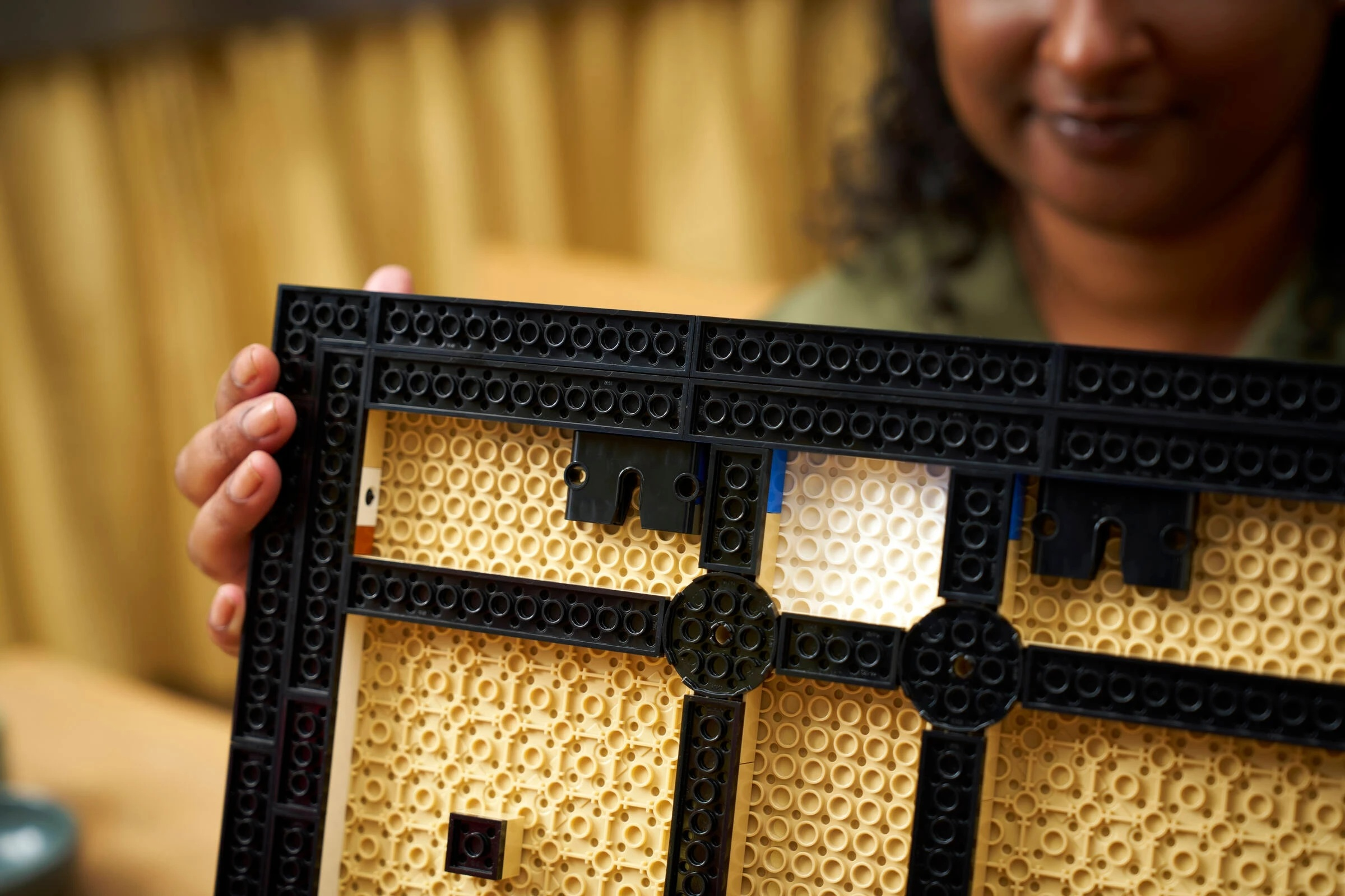 Ręka trzyma płytkę konstrukcyjną Lego z czarnymi i beżowymi klockami. W tle uśmiechnięta osoba.