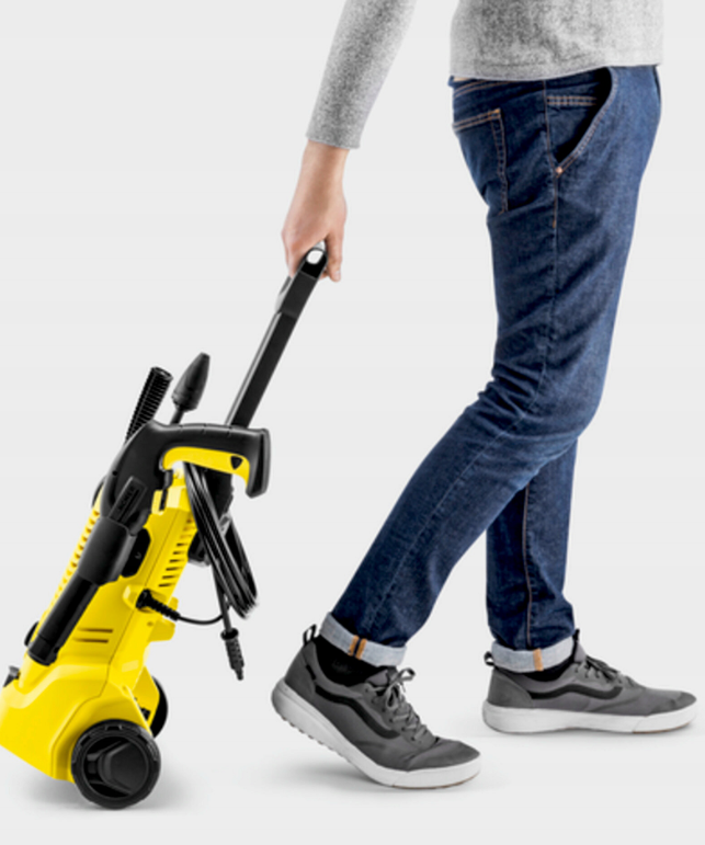 Osoba ciągnie żółtą myjkę ciśnieniową z kółkami i czarnym uchwytem.