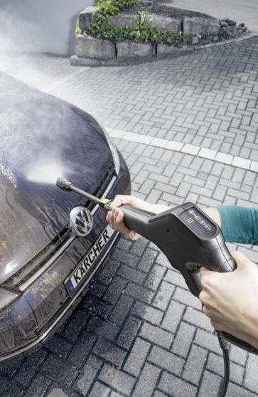 Osoba myje ciemny samochód myjką ciśnieniową na wybrukowanym podjeździe.