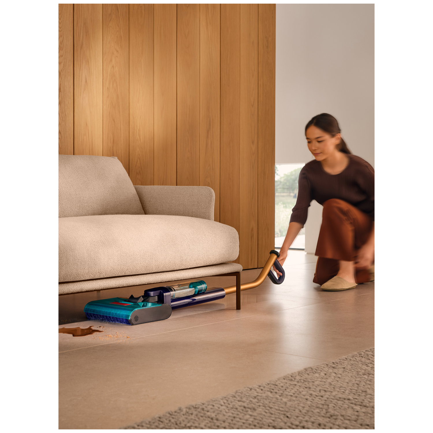 A woman vacuums under a sofa. A blue-headed vacuum cleaner is on the floor.