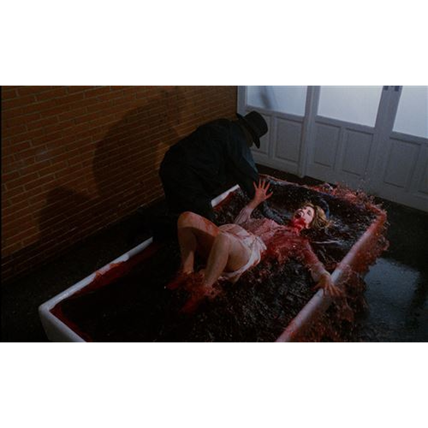 Une femme en chemise blanche gît dans une baignoire de liquide rouge, un homme en noir se tient à côté.