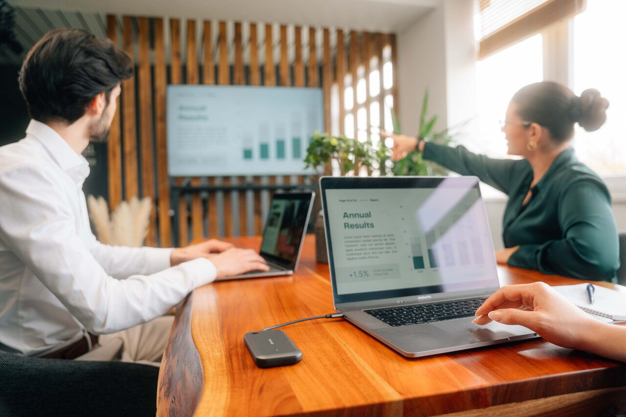 Ludzie pracują na laptopach przy drewnianym stole. Ekran pokazuje wykresy.