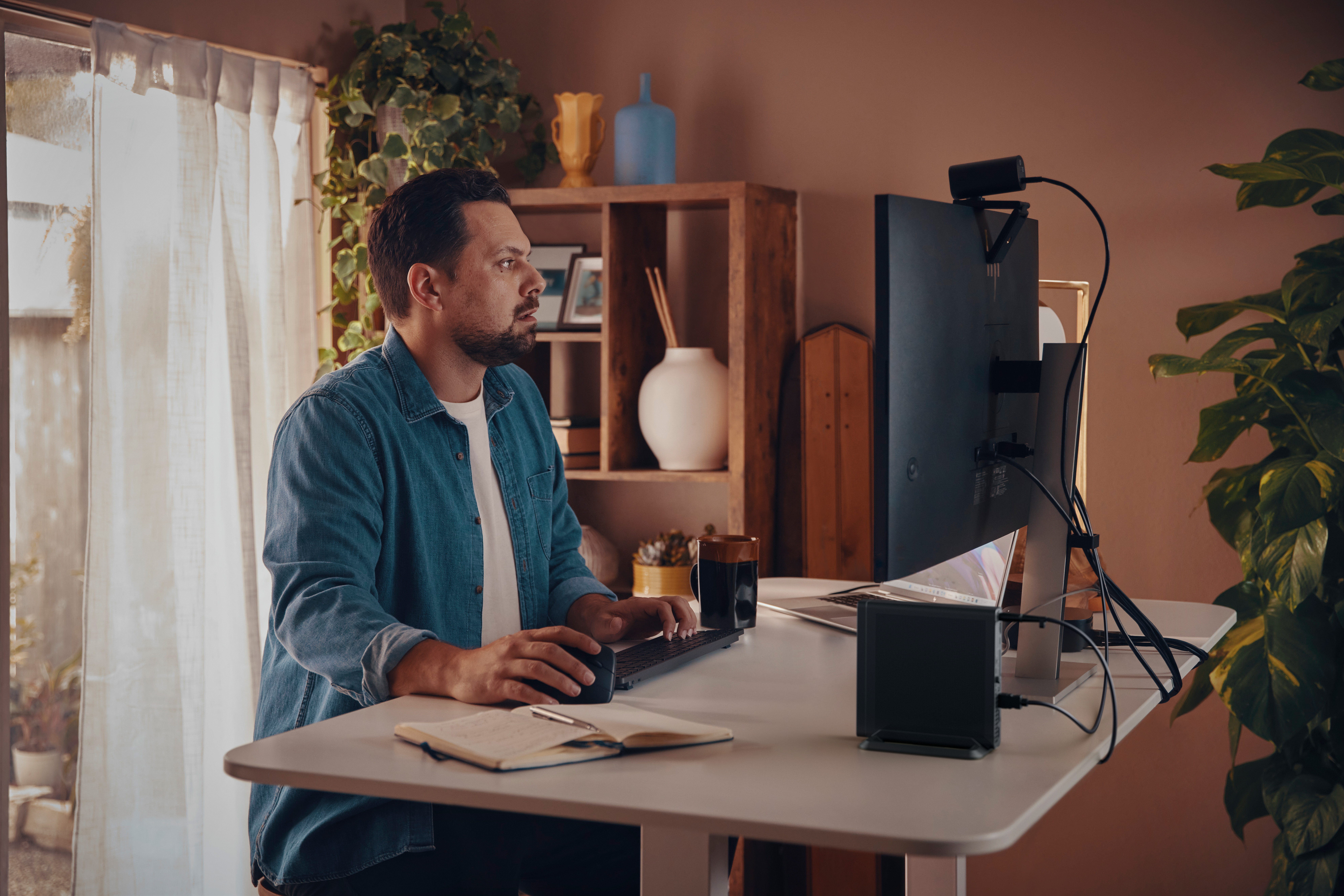 Mann in Jeanshemd arbeitet am Schreibtisch mit Computermonitor und Laptop.