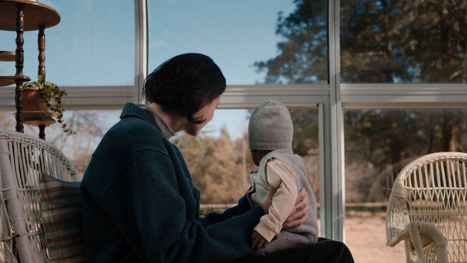 Frau hält Baby in einem Sessel vor einem großen Fenster.