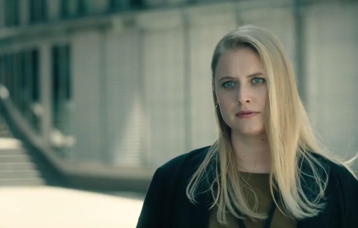 Eine Frau mit blonden Haaren und blauen Augen steht vor einer grauen Wand.