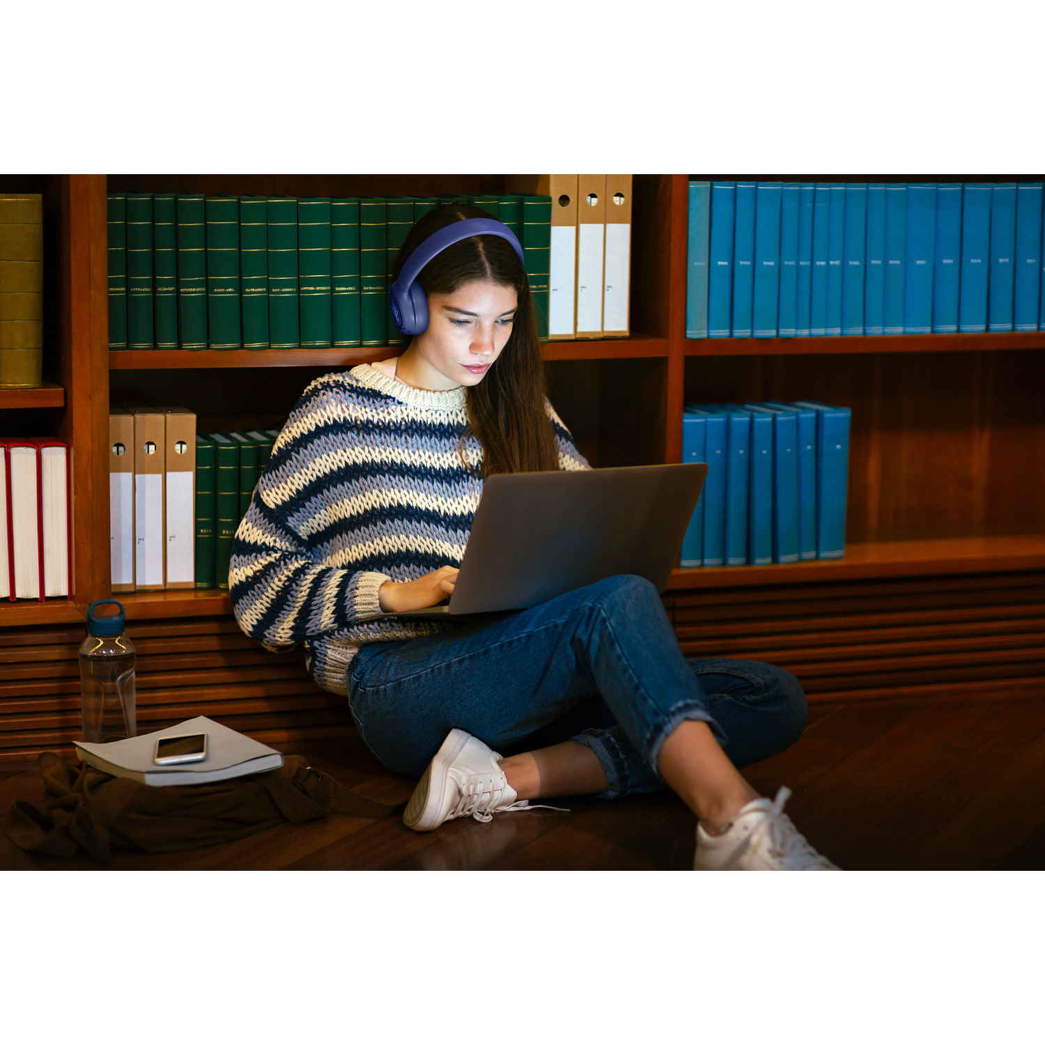 A young woman wearing headphones sits on the floor using a laptop.