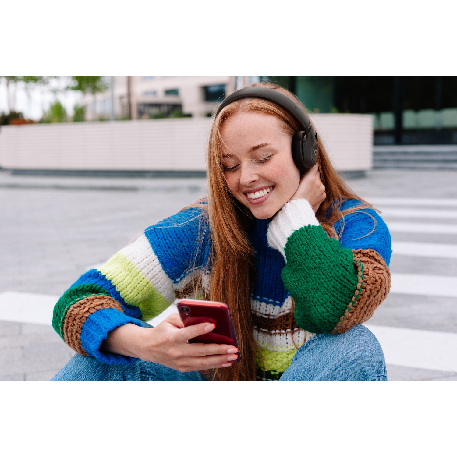 Une rousse avec un casque et un pull coloré sourit en regardant son téléphone.