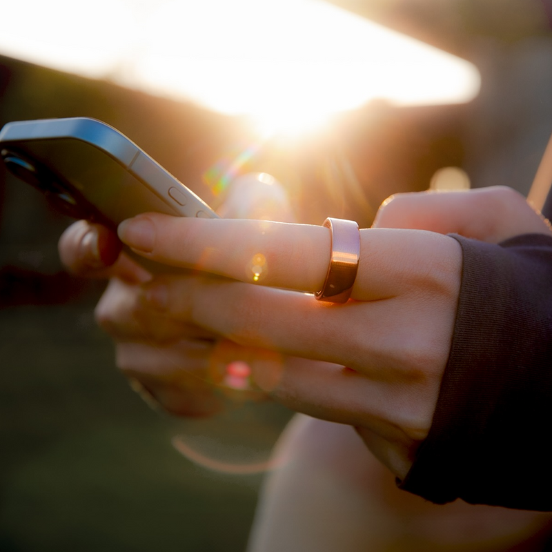 Közelkép telefonokat tartó kezekről, egy gyűrűvel az egyik ujjon. Napfény-törések.
