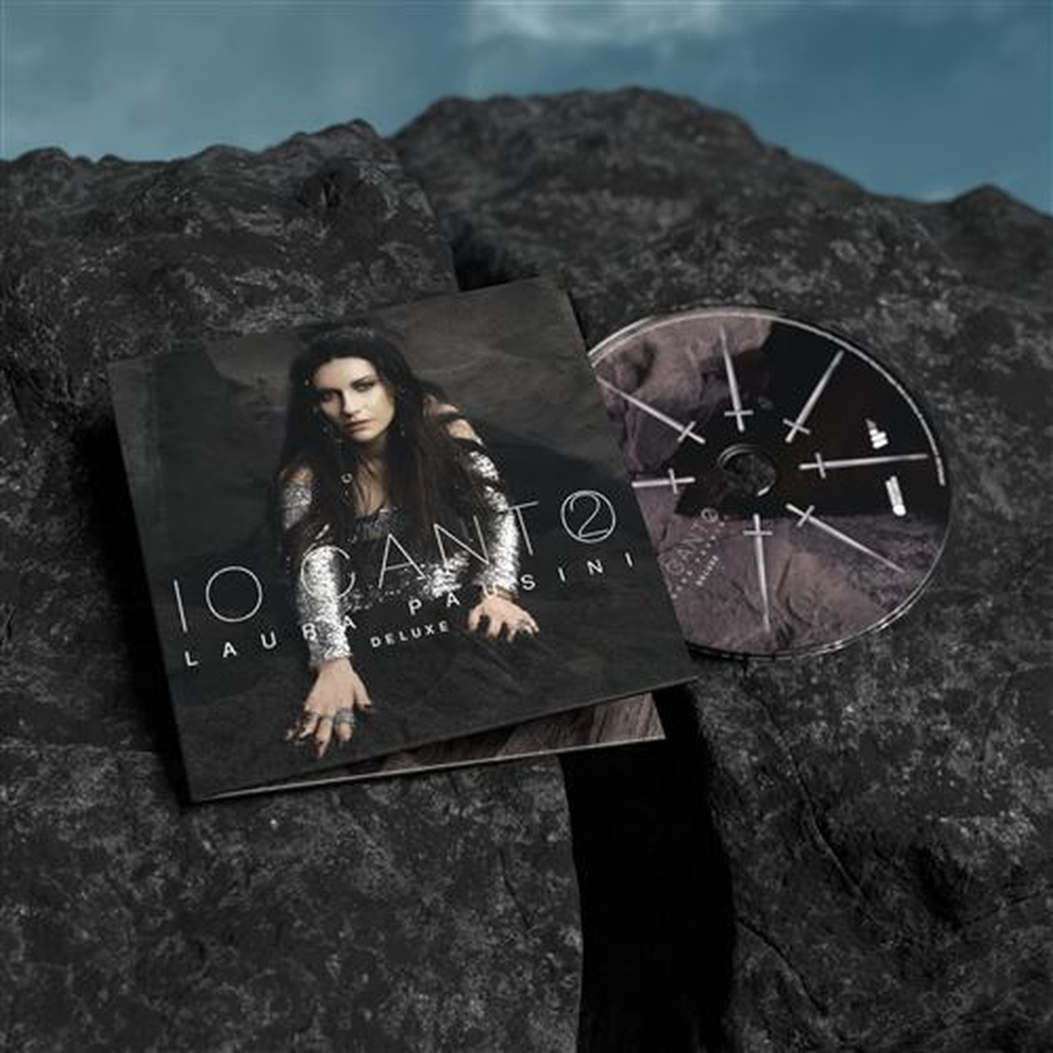 CD en hoes van Laura Pausini's "IO CANTO 2" op donkere rotsen onder bewolkte hemel.